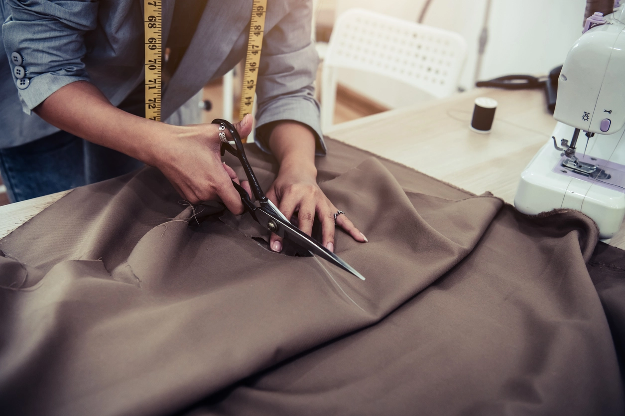 A person cuts brown fabric with scissors on a table, with a sewing machine, thread, and measuring tape nearby.