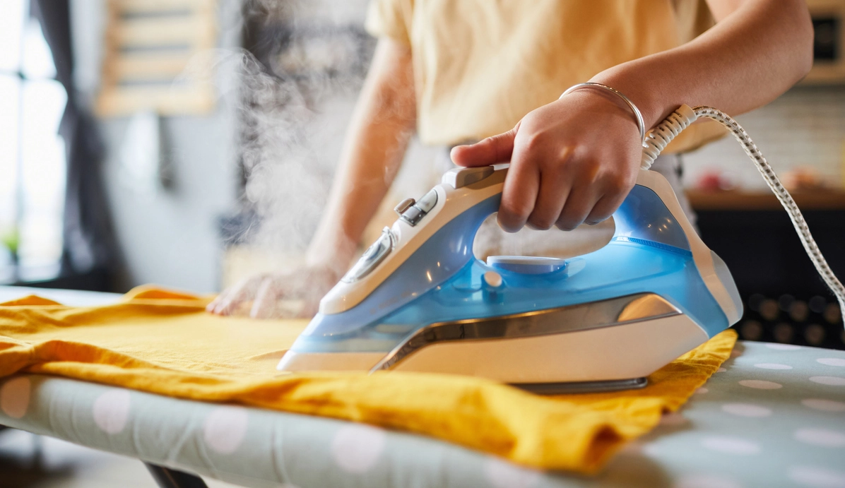 A person ironing a yellow shirt with a blue and white steam iron on an ironing board.