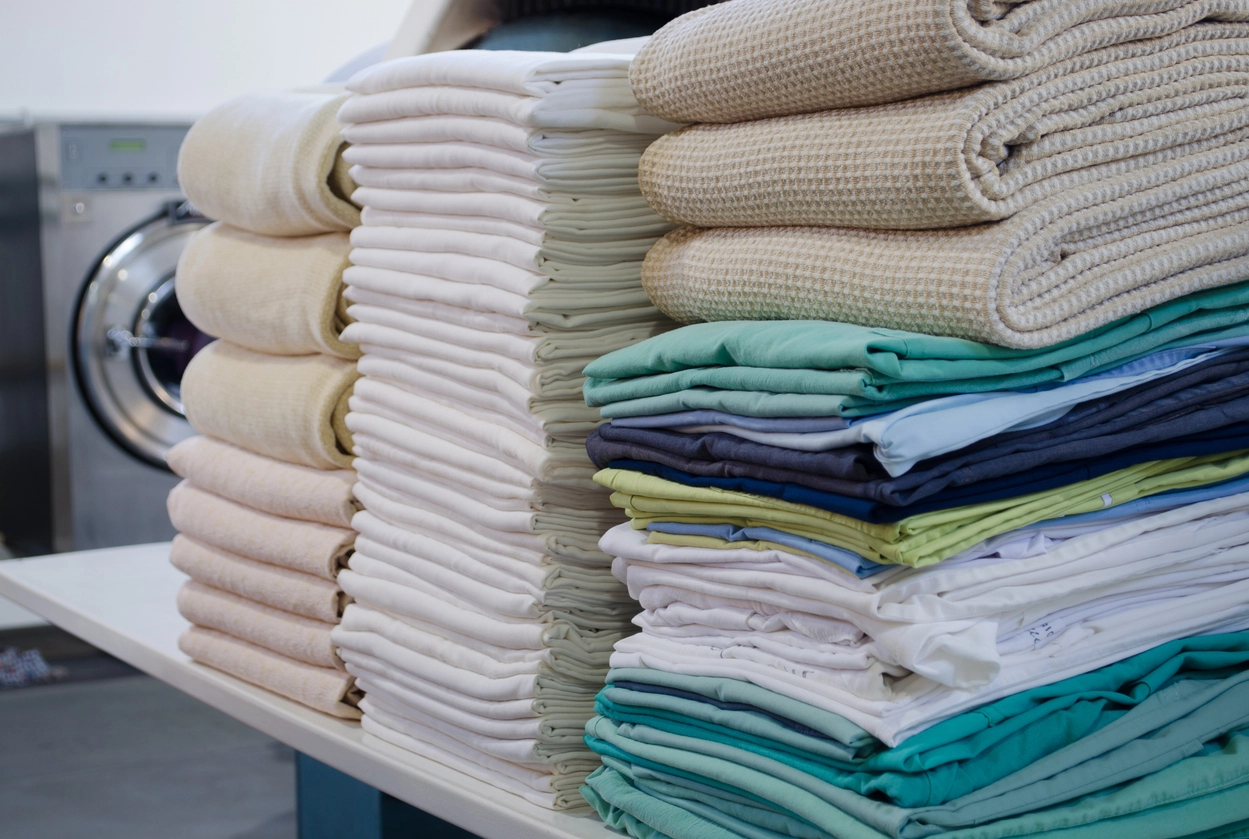 Stacks of neatly folded towels and linens in various colors on a table, with a washing machine visible in the background.