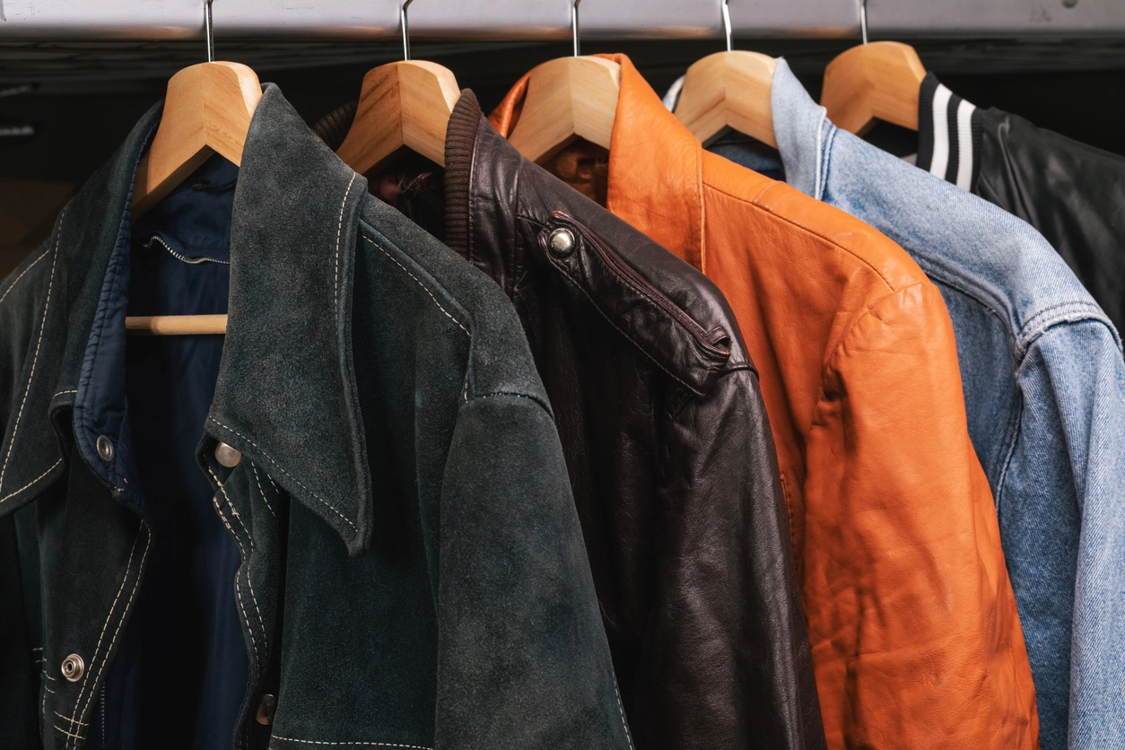 Five jackets of various materials and colors, including suede, leather, and denim, hang on wooden hangers on a clothing rack.