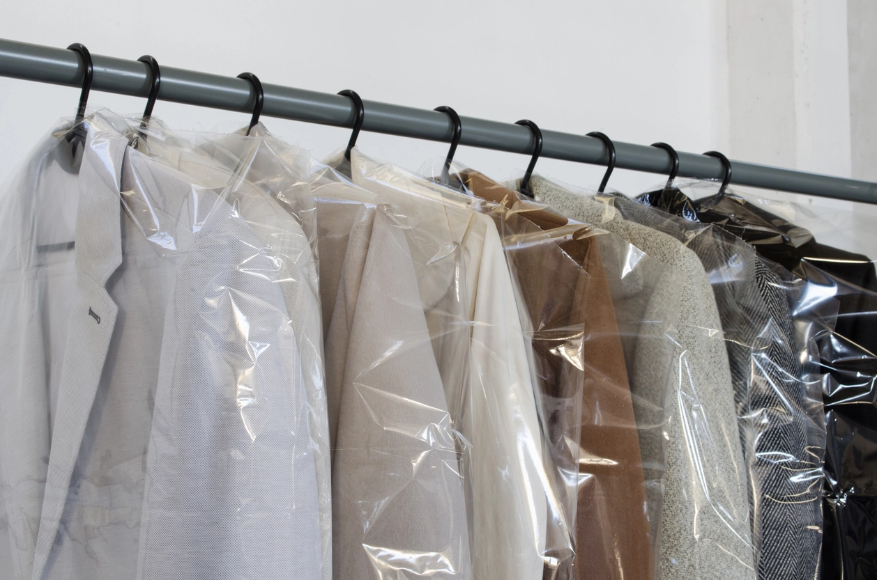 A row of assorted clothing items in neutral colors hang on black hangers, each covered with a clear protective plastic garment bag.