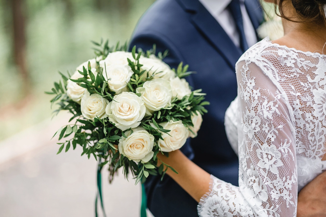 Close-up of a bride holding a bouquet of white roses and greenery, standing with a groom in a navy suit outdoors.