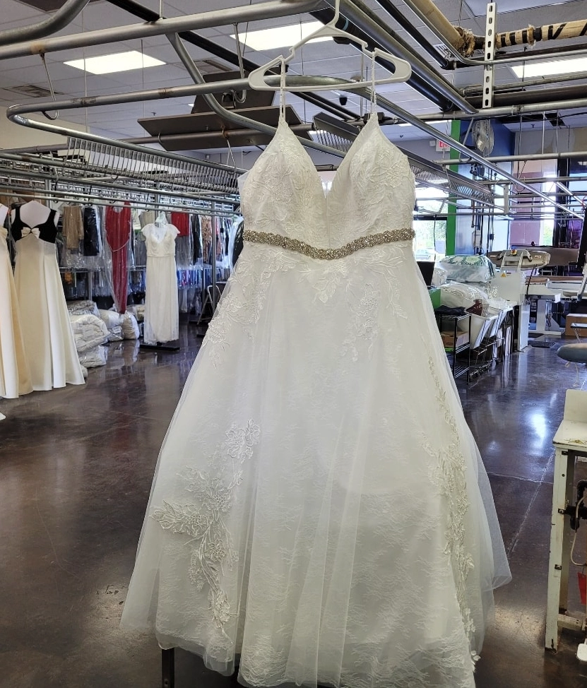 A white lace wedding dress with a beaded waistband hangs in a dry cleaning or alteration shop, surrounded by other garments and equipment.