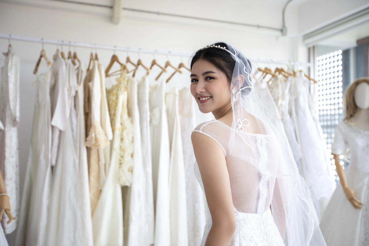 A woman in a white wedding dress and veil smiles while standing in front of a rack of hanging dresses in a bridal shop.
