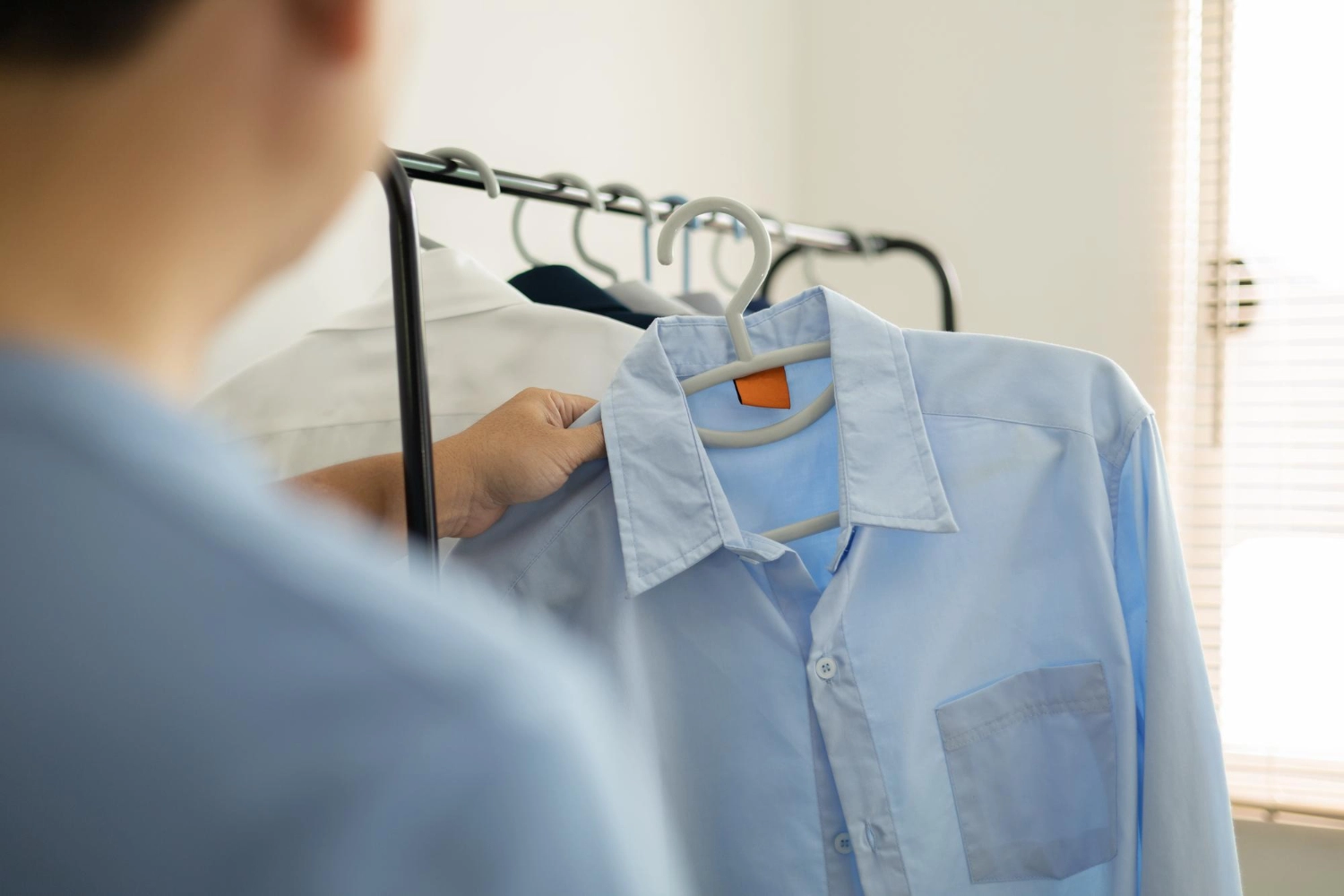 A person stands in front of a clothing rack, holding a light blue button-up shirt on a hanger.