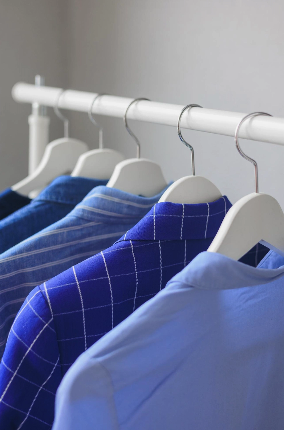 Five blue shirts in different shades and patterns hang neatly on white hangers on a white clothing rack.