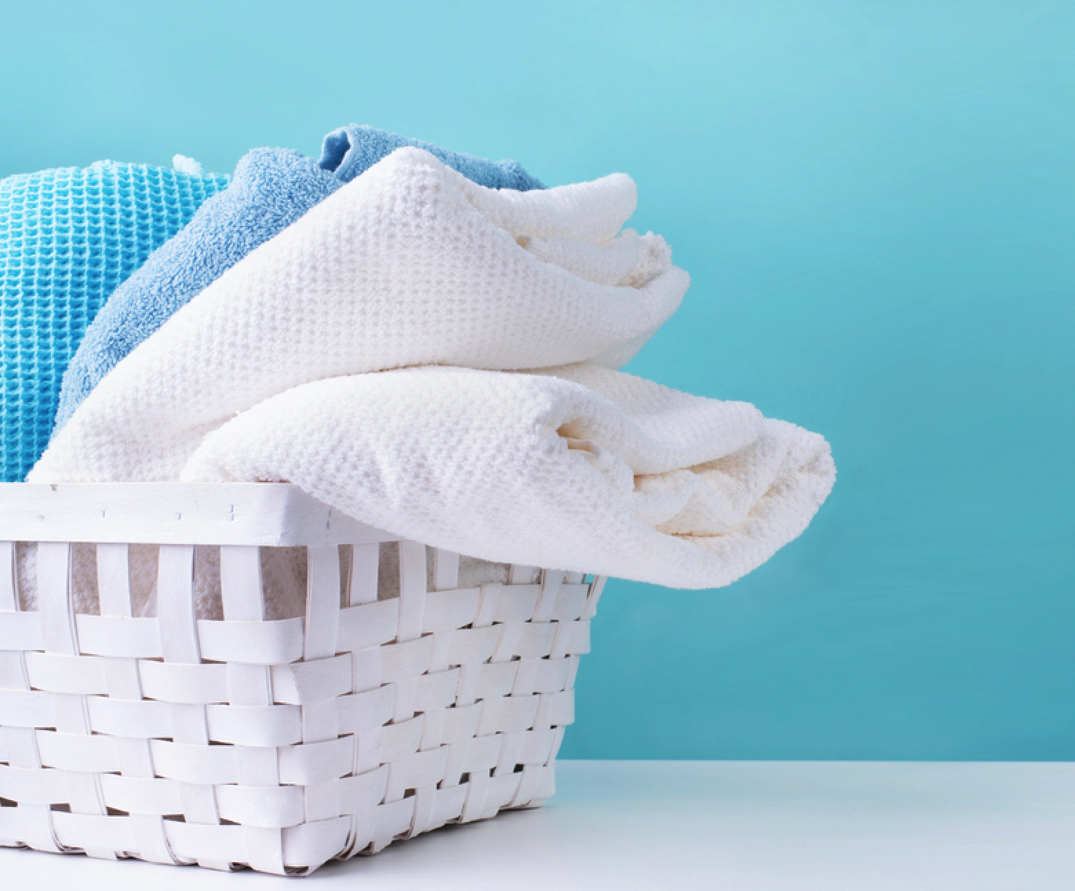 A white woven basket filled with folded blue and white towels sits on a white surface against a light blue background.