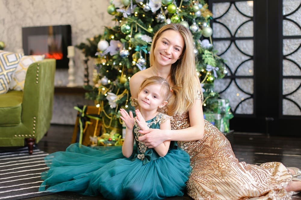 A woman and a young girl in formal dresses sit together in front of a decorated Christmas tree, smiling at the camera.