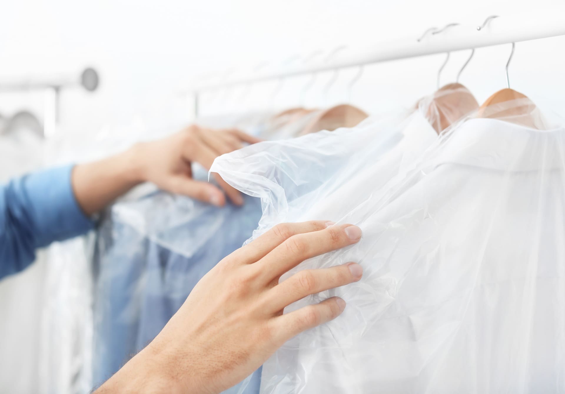 A person arranges freshly dry-cleaned shirts on hangers, each covered in clear plastic garment bags, on a white clothing rack.