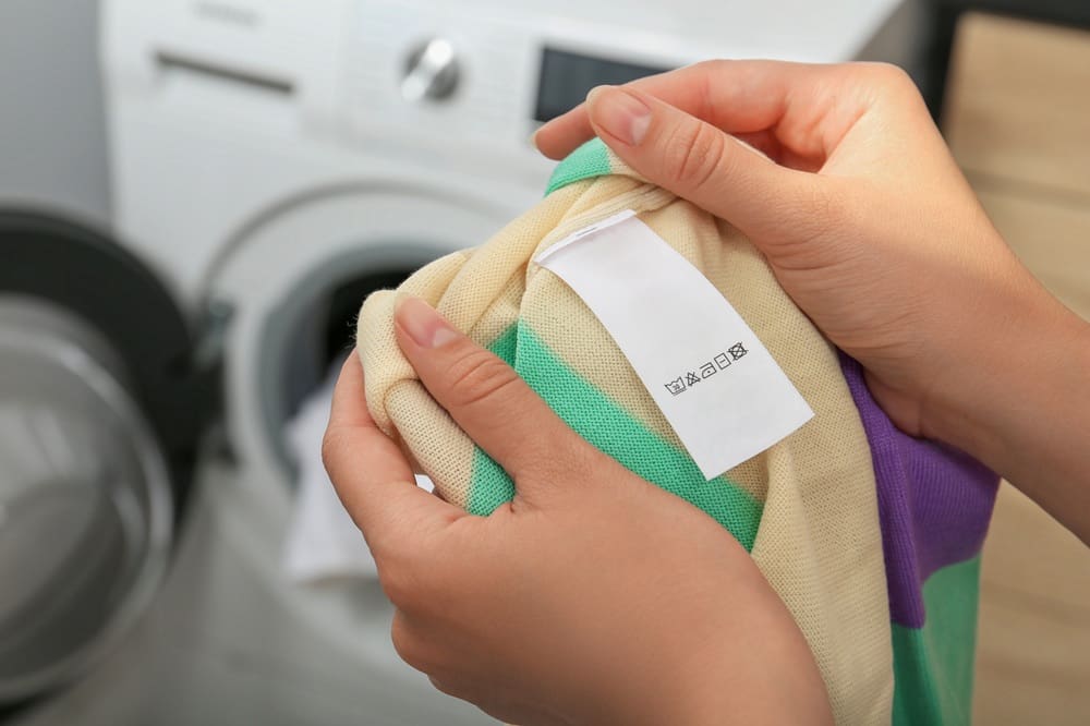 Hands holding a piece of clothing with a care label showing laundry symbols, in front of a washing machine.