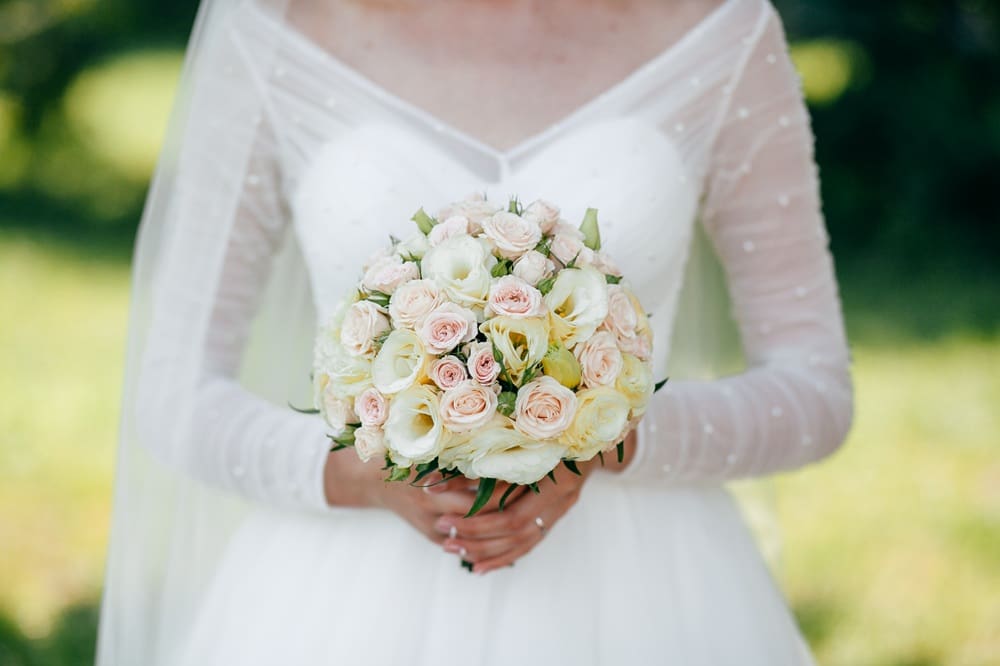 A bride in a white dress holds a bouquet of white and pale pink roses, standing outdoors with greenery in the background.