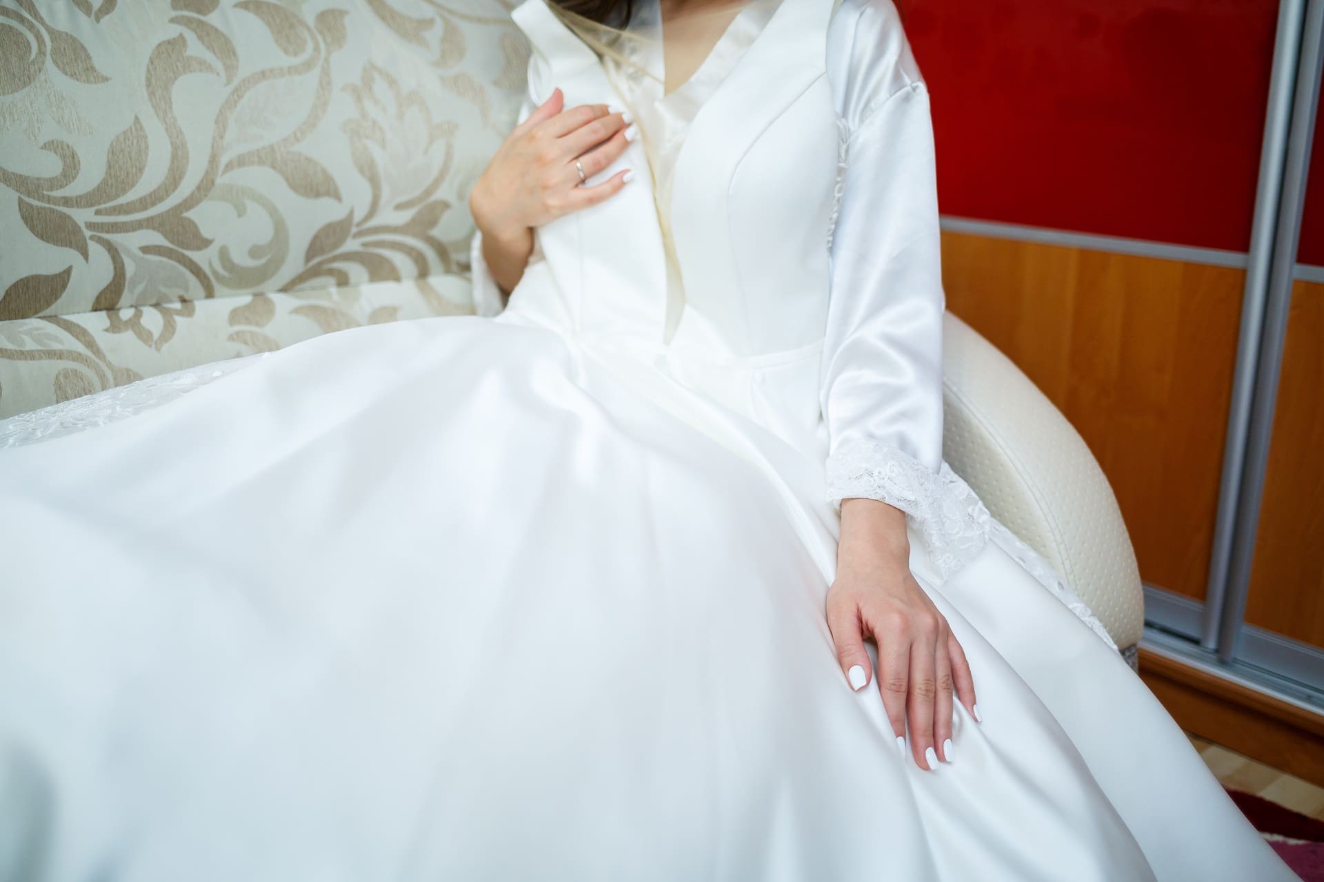 Person in a white wedding dress sits on a patterned sofa, with hands resting on their lap and dress.