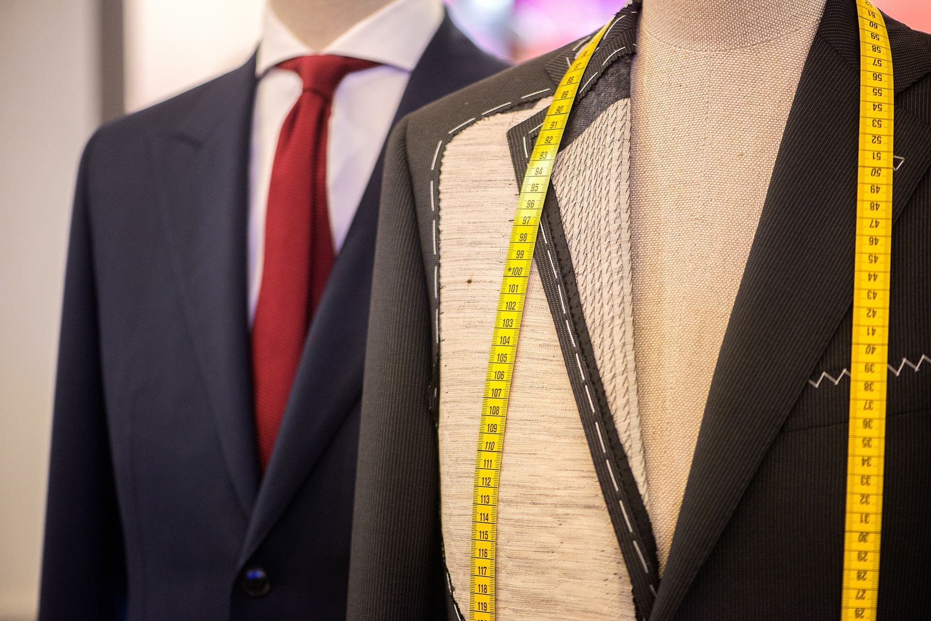 Two mannequins display a finished navy suit with a red tie and a partially constructed pinstripe jacket with a yellow measuring tape draped over the shoulders.