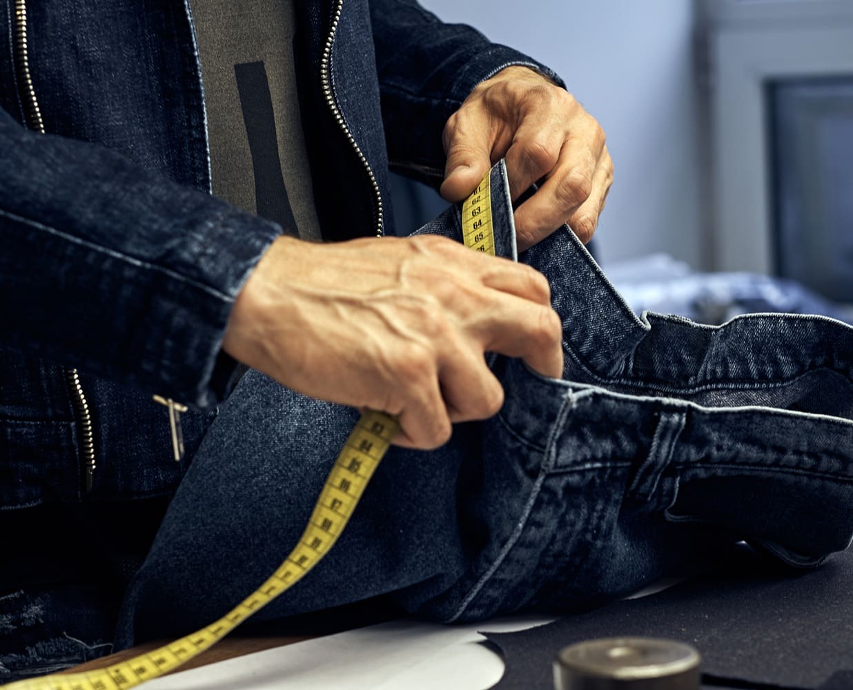 Person measuring a pair of dark blue jeans with a yellow tape measure on a work surface; only hands and torso are visible.