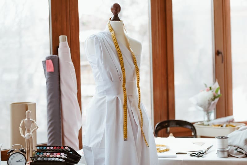 A dress form draped with white fabric and a yellow measuring tape stands in a well-lit sewing studio, surrounded by sewing supplies and tools.