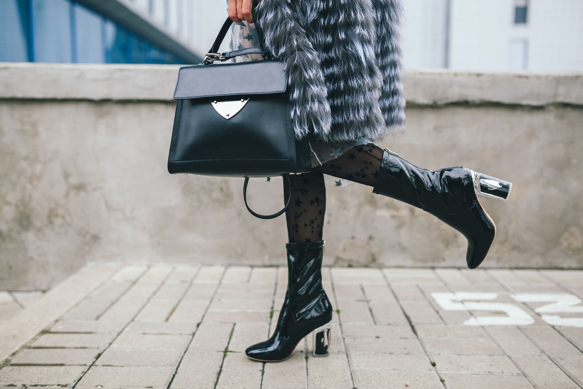 Person wearing a fur coat, patterned tights, and shiny black heeled boots, holding a black handbag while standing on a paved surface.