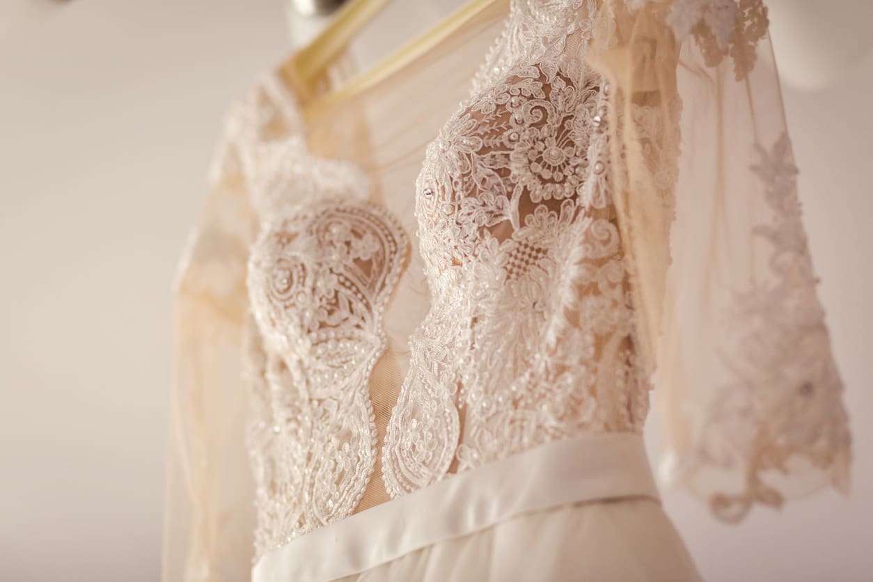 Close-up of an ornate lace wedding dress with sheer sleeves and intricate embroidery, hanging on a hanger in soft lighting.