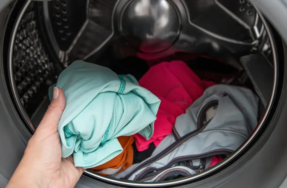 A hand places a folded piece of light blue clothing into a washing machine filled with various colored clothes.