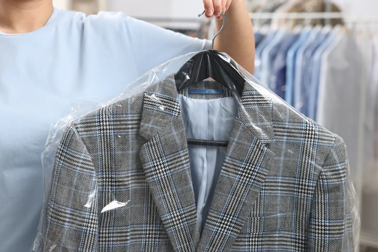Person holding a hanger with a plaid suit jacket covered in plastic, with other clothing items hanging in the background.
